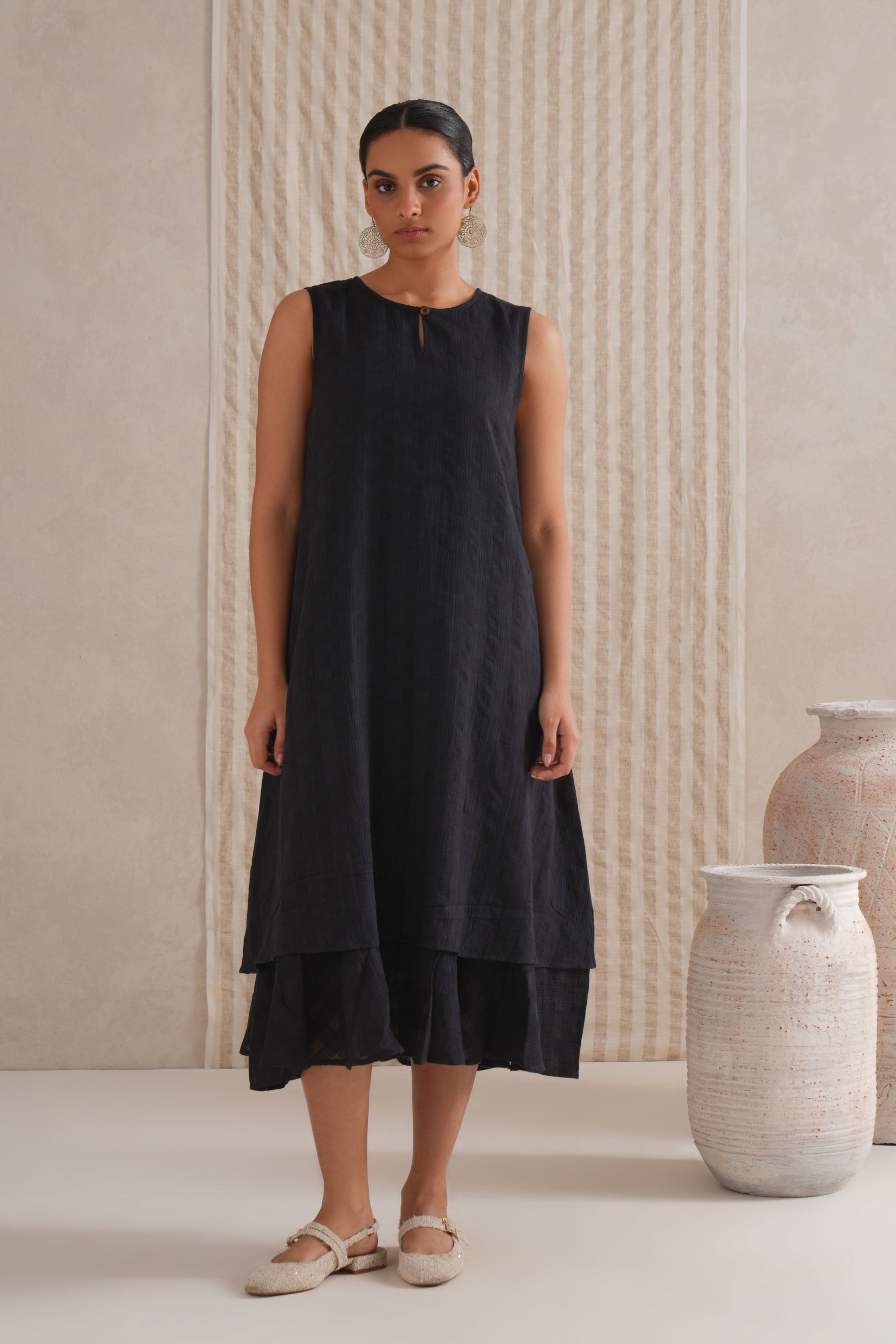 Woman wearing a black sleeveless dress standing in front of textured walls and vases.